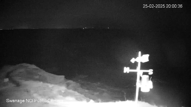 A dark scene showing a body of water under low light conditions, possibly at night. In the foreground, a weather vane is visible, indicating directions with the letters N, S, E, and W. The surrounding area is mostly shadowy with indistinct landforms in the background. A timestamp in the upper right corner shows "25-02-2025 20:00:36."