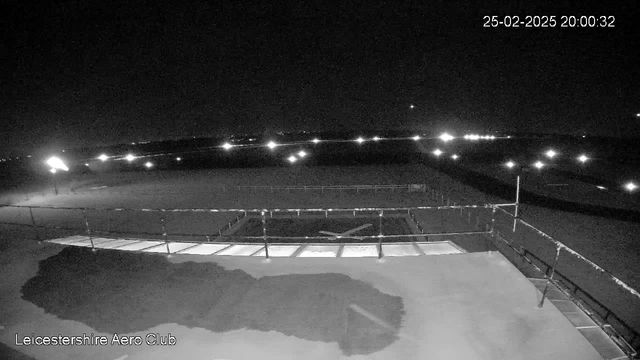 A nighttime webcam view of a dark landscape at Leicestershire Aero Club. The image shows a flat area with a lighted runway or tarmac, illuminated by scattered lights in the distance. There is a railing visible in the foreground, with shadows cast on the ground. The sky is mostly dark with some distant lights twinkling. The timestamp reads 20:00:32 on February 25, 2025.