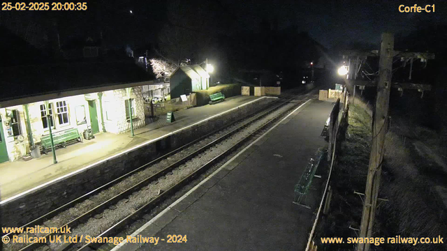 A nighttime view of Corfe Castle railway station. The station features a stone building with green accents, a row of benches, and a wooden fence. There are two railway tracks visible, with gravel in between. A green sign reads "WAY OUT." There is a light illuminating the area, and a wooden structure and trees can be seen in the background. The sky is dark with a hint of brightness.