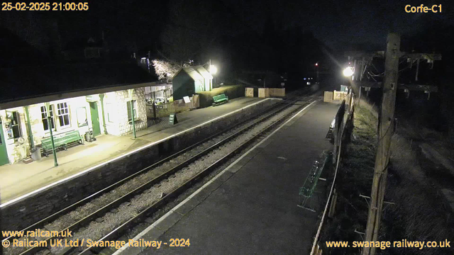 A dimly lit train station at night, featuring two parallel train tracks. On the left, there is a stone building with large windows, a green bench, and a light above. To the right, there are more benches and a wooden fence marking an exit area. A sign is visible indicating a way out. The scene is devoid of people, and the surrounding area is dark.