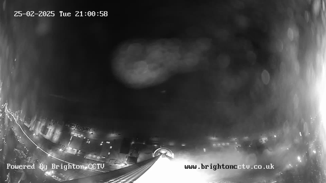 A dark nighttime scene captured from a webcam, showing a cityscape with faint lights from buildings and streets below. The image is predominantly black with lightened areas indicating illuminated structures. A possible mist or water droplets affect the clarity, creating a blurred effect in parts of the image. The date and time at the top indicate it is February 25, 2025, at 21:00:58. A watermark at the bottom displays "Powered By Brighton CCTV" alongside a website URL.