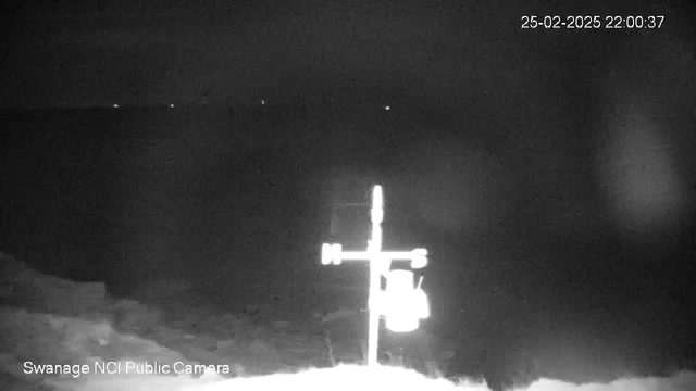 A dimly lit webcam image showing a blurred view of the sea at night. In the foreground, a white structure resembling a weather vane stands with directional markings for North, South, East, and West. The background is mostly dark, with faint lights visible on the horizon, suggesting distant boats or land. The date and time display in the top right corner indicates it is February 25, 2025, at 10:00 PM.