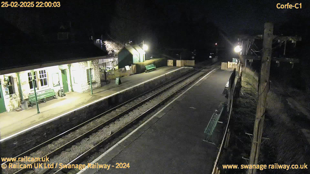 A dimly lit railway station at night. The platform features stone walls and green waiting benches. On the left, there is a building with large windows, illuminated from within. A sign that reads "WAY OUT" is placed on the platform. Two sets of railway tracks run parallel in the foreground, with a wooden fence and shrubbery visible on the right side of the image. In the background, a distant light source suggests the presence of another building or area. The scene is tranquil and empty, evoking a sense of quiet solitude.