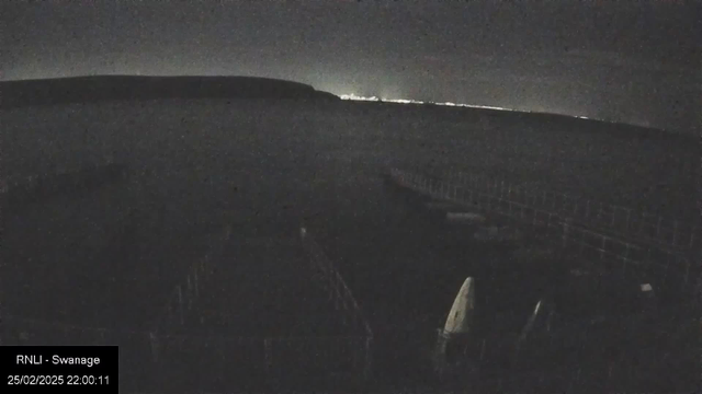 The image shows a dimly lit scene of a coastline at night. The horizon is faintly visible in the distance, where a thin line of lights suggests a nearby town or city. In the foreground, a structure made of wooden beams can be seen, likely a pier or dock, with various shapes including boats or other objects sparsely placed along it. The overall tone of the image is dark, with shadows dominating the scene.