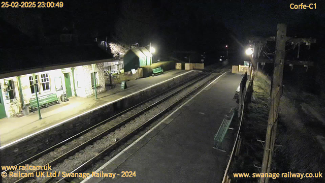 A quiet railway station at night, with platforms illuminated by a faint light. The stone building on the left features large windows and a covered waiting area with green benches. To the right, there is a wooden fence with a "Way Out" sign, and another green bench is visible. The platform is empty, with two railway tracks running parallel, bordered by dark surroundings. There are no trains present, and the scene conveys a tranquil nighttime atmosphere.