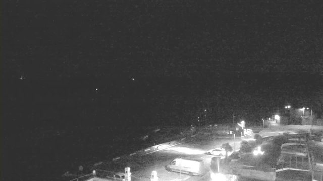 A dark scene captured by a webcam, showing a coastal area at night. The foreground features a partially illuminated parking lot with several parked vehicles, including a white van. In the background, the ocean appears mostly black with a few faint lights visible far off in the distance, suggesting the presence of boats or distant land. The overall atmosphere is quiet and dimly lit, with a few light sources creating shadows.