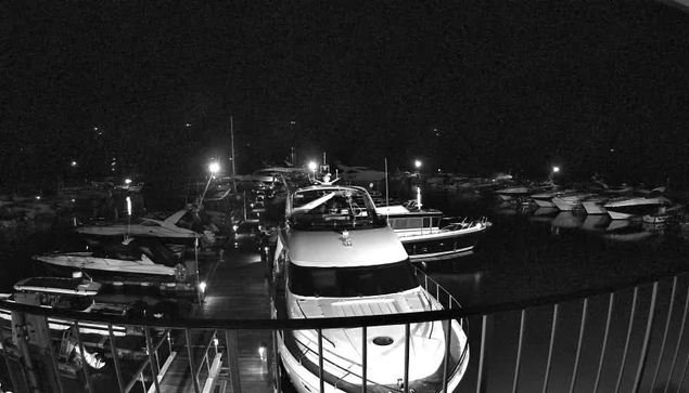 A black and white image of a marina at night, featuring several boats and yachts docked along a wooden pier. The water reflects the sparse lights from the boats and surrounding area, creating a calm, serene atmosphere. The scene is predominantly dark, with faint outlines of boats in the background. Some boats are illuminated with soft lights, while others are partially obscured by shadows.