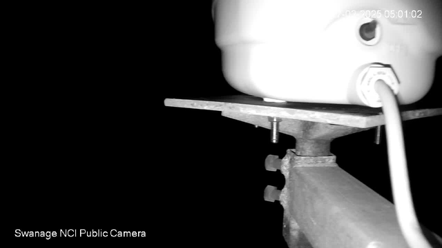 A close-up image of a white cylindrical object mounted on a metal platform, with visible screws and a power cord. The background is dark, creating high contrast, and faint text in the lower left corner reads "Swanage NCI Public Camera." The timestamp in the upper right indicates the date and time.