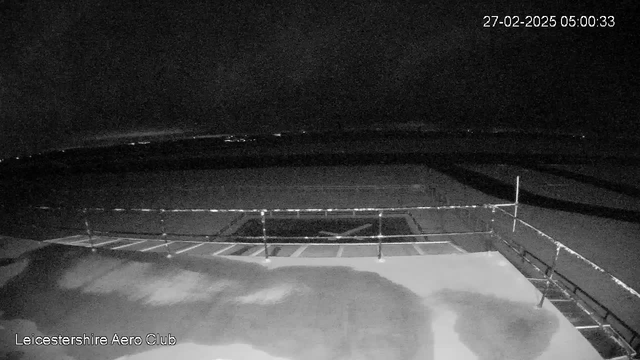 A dark scene captured from a webcam at Leicestershire Aero Club, showing a flat, open area with shadows and faint outlines of surfaces in the foreground. In the background, there are lights shimmering faintly in the distance against a dark sky. The time stamp in the corner indicates it is 5:00 AM on February 27, 2025.