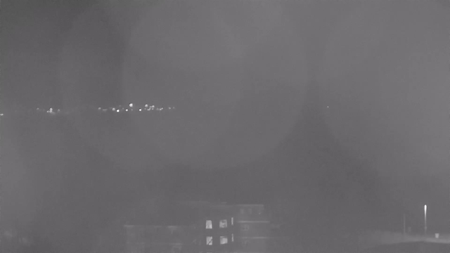 A dark, blurry night scene captured from a webcam. In the foreground, a structure with windows is partially visible, suggesting an urban environment. In the background, there are faint lights, possibly from distant buildings or streetlights, scattered across the horizon, surrounded by darkness. The overall image has a grayish tone, making details difficult to distinguish.