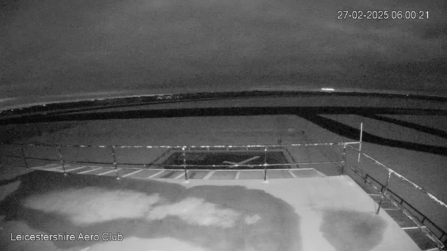 A dimly lit outdoor scene from a webcam showing a runway and a small section of the tarmac at Leicestershire Aero Club. The image is predominantly dark gray with a few lighter patches indicating snow or wet surfaces. A low railing borders the foreground. The sky is cloudy, with a faint glow on the horizon suggesting the presence of city lights in the distance. The date and time are displayed in the top right corner.