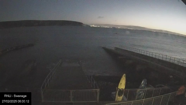 A peaceful early morning scene by the water. The sky is transitioning from dark to light, with hints of dawn on the horizon. The sea is calm and reflects the dim light. In the foreground, a small section of a metal railing surrounds a concrete pier. There are two kayaks, one yellow and one blue, resting against the railing. In the background, a distant coastline is visible under the softly illuminated sky.