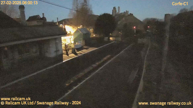 A dimly lit scene of a small railway station at dawn. The foreground shows empty train tracks leading into the distance. On the left, there is a low building with a roof and an awning, illuminated softly by warm lights. To the right, there are scattered trees and a steep hill with structures at the top, creating a sense of depth. The sky is a gradient of blue, indicating early morning light.