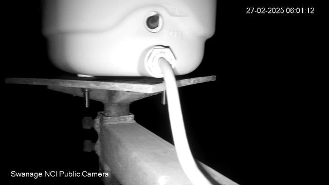 A close-up view of a white container resting on a metallic surface. The container has a nozzle and is connected to a cable. The background is mostly dark, indicating low light conditions. A timestamp in the upper right corner reads "27-02-2025 06:01:12." The lower left corner displays "Swanage NCI Public Camera."