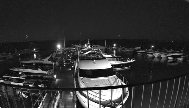 A nighttime view of a marina filled with multiple boats and yachts. The scene is predominantly dark, with bright artificial lights illuminating some boats and the dock. A large white yacht is positioned in the foreground, with its bow facing the camera. The water is still, reflecting some of the surrounding lights, while the silhouettes of additional boats and the dock are visible in the background.