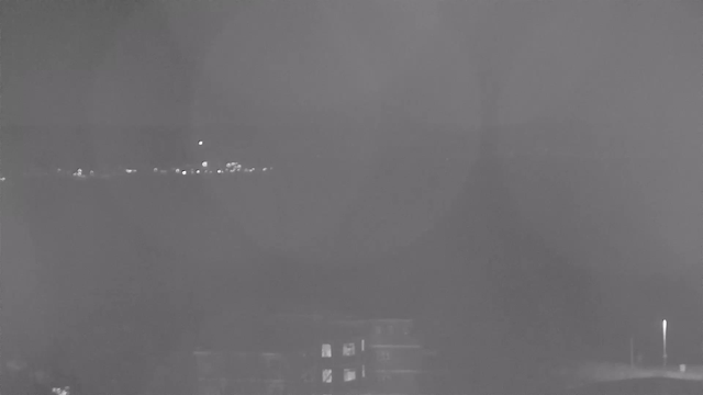 A dimly lit night scene captured by a webcam. In the foreground, there are dark silhouettes of buildings. In the background, faint lights are visible along a horizon, suggesting the presence of distant structures or a shoreline. The overall image is washed in grayscale, creating a somber atmosphere.