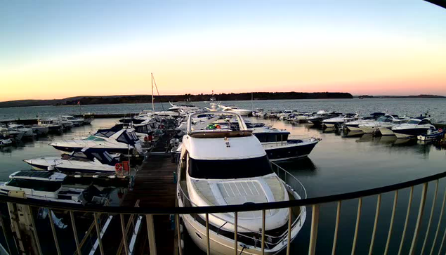 A marina filled with various boats and yachts docked along a wooden pier, reflecting in calm water. The sky transitions from soft orange to blue as the sun sets in the background, creating a serene atmosphere. A railing in the foreground frames the view, while distant hills are visible on the horizon.