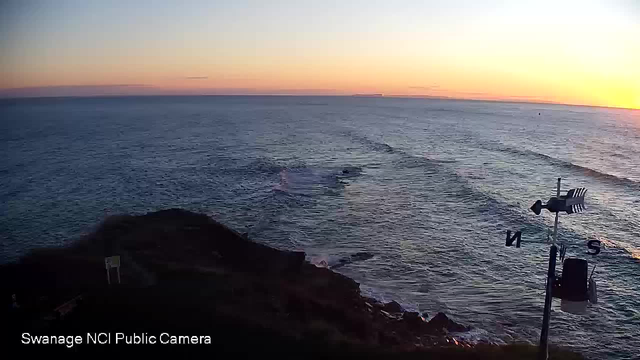 A coastal scene at sunset, with gentle waves lapping at rocky outcrops. The sky displays a gradient of colors ranging from orange to deep blue, indicating the transition from day to night. A weather vane is visible in the foreground, with cardinal directions marked. The horizon line meets the sea, and distant land can be seen in the background.