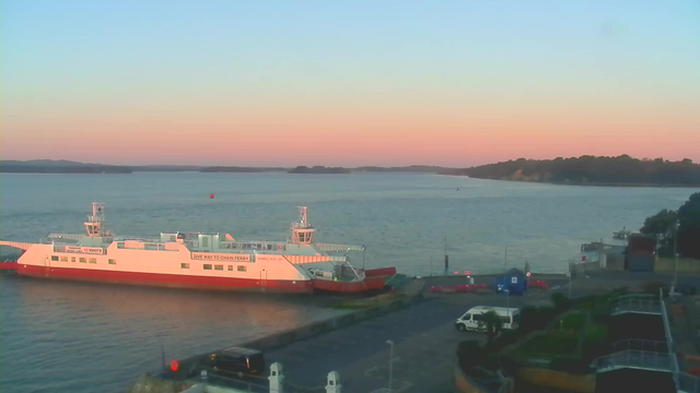 A calm waterfront scene during twilight, featuring a large white ferry with a red bottom docked near a shoreline. The sky transitions from soft pink to light blue, and the water reflects these colors. In the background, hills are visible along the horizon, while a small boat moves on the water. Various vehicles are parked near the dock, and there are dark green shrubs and a pathway in the foreground.