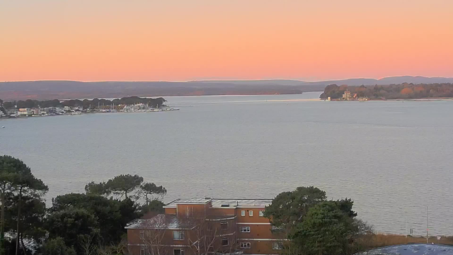 A panoramic view of a serene body of water during twilight. The sky transitions from soft orange to light purple as the sun sets. In the foreground, there are trees and a low building with a flat roof. To the left, a small town is visible along the shoreline with buildings and boats, while in the distance, a small island with structures can be seen. The water appears calm, reflecting the colors of the sunset.