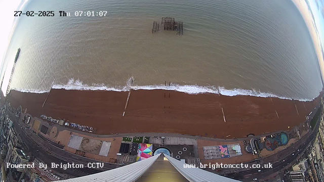 A bird's eye view of a beach with brown sand and gentle waves lapping at the shoreline. In the distance, there is a partially submerged pier structure in the water. The top of the image shows a clear sky, while the bottom displays a promenade with colorful attractions, pathways, and potential seating areas visible. The scene is tranquil, with minimal activity on the beach and promenade. A timestamp reads "27-02-2025, Thursday, 07:01:07."