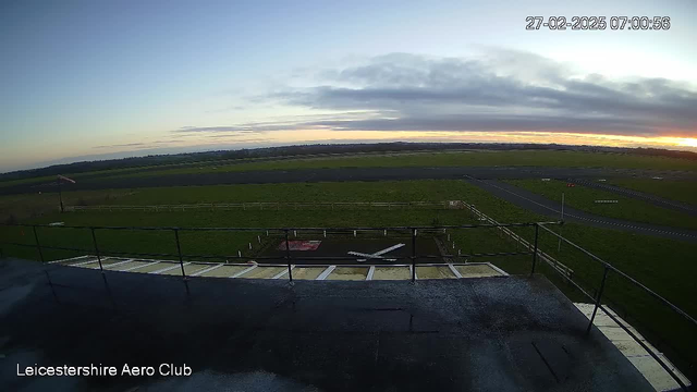 A webcam view from the Leicestershire Aero Club captures an early morning scene. The foreground includes a rooftop platform with a railing, leading to a grassy area with a small white airplane. A runway extends into the distance on the right, bordered by fencing. The sky features a gradient of colors from light blue to soft orange near the horizon, with some clouds scattered across the sky.