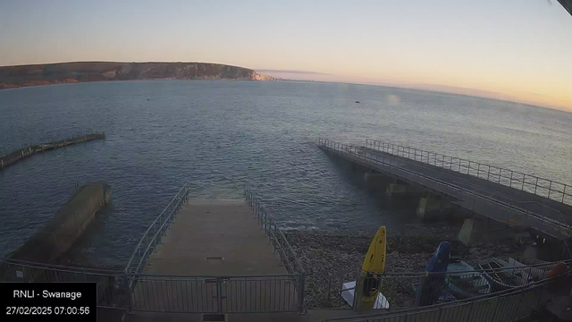 A coastal scene during early morning light shows calm waters with gentle ripples. In the foreground, there is a wooden pier leading into the water, flanked by a railing. To the left, a circular, smaller pier extends into the sea. On the right side, there are boats on the ground, partially covered, and a yellow kayak is visible leaning against a railing. The shoreline is rocky, and the backdrop features a distant cliff with subtle colors from the rising sun. The sky transitions from light blue near the water to soft hues of orange and pink towards the horizon.