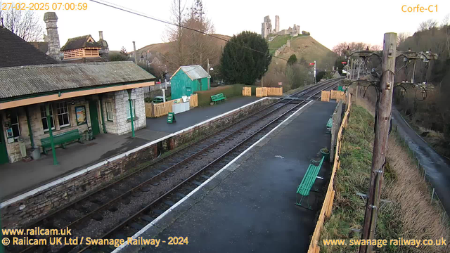 A railway station platform is visible, featuring green benches along the edge. In the background, a small green building and wooden fencing can be seen. A hillside rises behind the station, topped with a castle ruin. The scene is set in early morning light with clear skies and a few trees around the area. Electric posts and wires are present to the right, alongside railway tracks leading away from the platform.