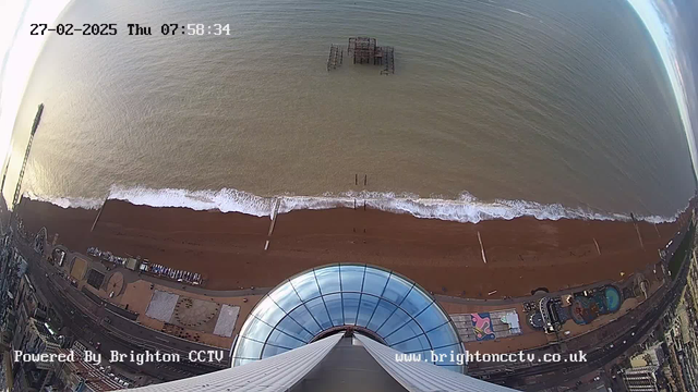 A high-angle view of a coastline shows sand and waves meeting the shore. In the background, the calm sea stretches to the horizon. A small, dilapidated pier stands in the water, and several boats are visible near the beach. The foreground features a large dome structure, likely part of an observation tower. The date and time appear at the top of the image, indicating it is early morning, and a logo for Brighton CCTV is in the lower part of the image.