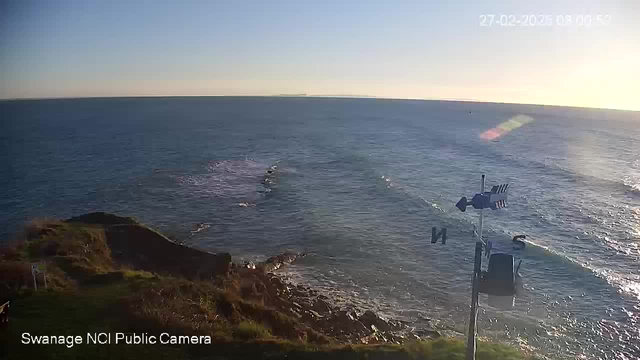 A coastal scene showing the ocean under a clear blue sky with the sun rising on the horizon. In the foreground, there is grassy land with rocks on the shoreline. To the right, a weather vane is visible, indicating the direction of the wind. The water has gentle waves, and there are small ripples sparkling in the sunlight.