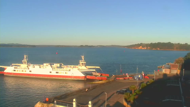 A ferry is docked at a harbor on a calm body of water. The ferry is predominantly white with orange details and has various signs and equipment visible. In the background, there are green hills and trees lining the shore. The sky is clear and blue, indicating a sunny day. There are a few structures near the water's edge, including a red building and some parked cars. The setting is peaceful, with gentle water ripples and distant land visible on the horizon.