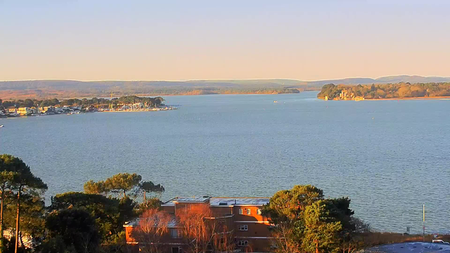 A panoramic view of a calm body of water, bordered by a shoreline with trees and buildings. In the distance, small boats can be seen on the water, and across the bay, there are more trees and hills. The sky is clear, with a soft light suggesting early morning or late afternoon. The foreground features trees and a low building, indicating a rural or coastal area.