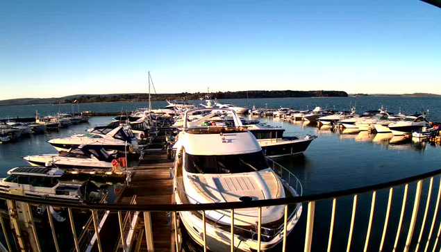 A sunny waterfront scene featuring a marina filled with multiple boats and yachts docked at the pier. The water is calm and reflects the blue sky. In the background, there are green hills and trees lining the shore. The perspective is from a high vantage point, showcasing the arrangement of the boats and the serene surroundings.