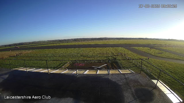 A wide outdoor view from a high vantage point at Leicestershire Aero Club. The image captures a clear blue sky with a few clouds, and a green grassy area below. In the foreground, there is a flat surface with a railing, and a section of the ground appears to have a white outline resembling an airplane. The background shows an expansive field with a runway and a few scattered trees in the distance. The setting is bright and sunny, indicating early morning light.