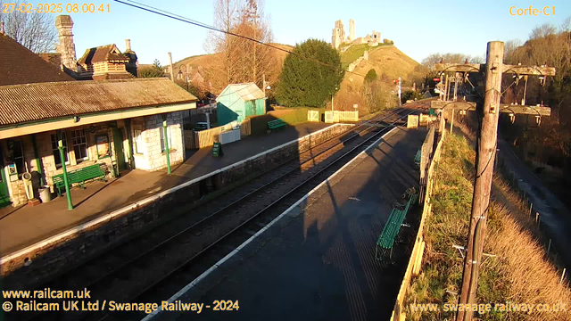 A view of a train station in the early morning light. The foreground shows a platform with several green benches and a sign indicating "WAY OUT." To the left, there is a small building with stone walls and a sloped roof. The background features a hill with a castle ruin and trees. A railway track runs parallel to the platform, and wooden utility poles with wires are visible on the right. The sky is clear and bright.