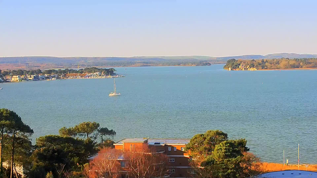 A wide view of a calm body of water, likely a bay or estuary, under a clear blue sky. In the foreground, there are some buildings surrounded by trees. A small white sailboat is visible on the water, with a marina and a line of buildings along the shore in the distance. Beyond the water, there are gentle hills or low mountains on the horizon.