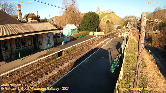 A sunny morning scene at a train station with a clear blue sky. In the foreground, there are two railway tracks running parallel, bordered by a low stone wall. To the left, a traditional stone station building features a sloped roof and columns. There are green benches on the platform and a green sign that reads "OUT." In the background, a tall hill rises, topped by ruins of a castle, surrounded by trees. A wooden telephone pole with wires stands on the right side of the image.