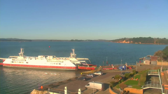 A large white ferry with red accents is docked at a pier, beside calm blue water. The scene is set under a clear blue sky. In the background, green trees line a shore that rises into gentle hills. Near the ferry, there are a few parked cars and a small building. The area is well-maintained, with a grassy section and benches visible.