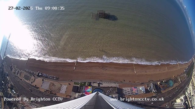 Aerial view of a beach with sandy shoreline and gentle waves lapping at the shore. The water appears calm, reflecting sunlight. In the distance, a derelict pier structure is visible in the water. The beach is lined with various structures, including colorful tents and a pool area. People can be seen walking along the beach. The image is timestamped with the date and time at the top left corner, and a logo at the bottom left.