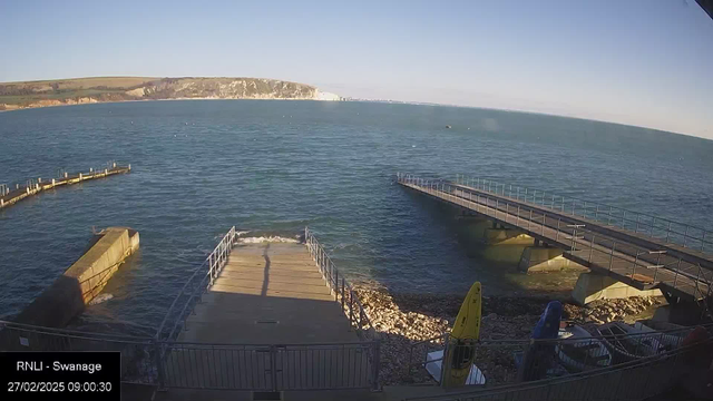 A coastal scene depicting calm blue waters under a clear sky. In the foreground, there are two piers extending into the water: one is made of wood and is slightly curved, while the other appears more straight with a railing along its sides. There are several boats along the shore, mostly covered, with one bright yellow kayak visible on the right. The shoreline is rocky, with small waves lapping against the piers. In the background, cliffs rise gently above the water. The image captures a peaceful, sunny day by the sea.