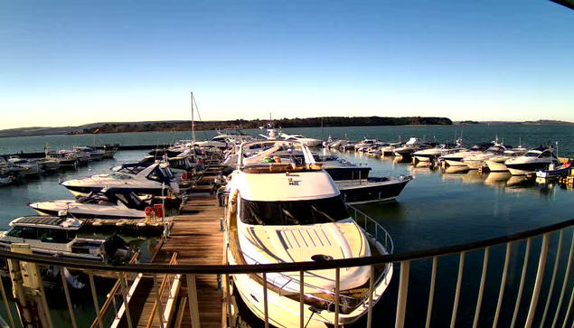 A marina scene with numerous boats docked in calm water, reflecting the clear blue sky. The image captures a variety of yachts and smaller vessels lined up along wooden docks. In the background, there is a green hill under the bright sky, enhancing the peaceful coastal atmosphere. The scene is bathed in natural light, indicating a sunny day.