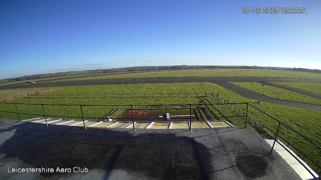 A clear blue sky is visible over a green field with a runway in the foreground. There are patches of grass and a fenced area. In the distance, low hills can be seen. The image is taken from an elevated position, showing a flat surface in the lower part with steps leading down. The webcam timestamp indicates the date and time.