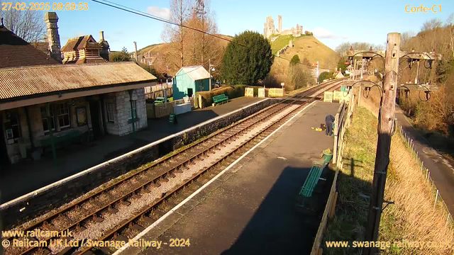 A sunny day at Corfe Castle train station, featuring a broad view of the platform and railway tracks. On the left, a historic stone station building with a chimney, partially covered in shadow. Green benches line the platform, and a blue shed is visible in the background. A person is bending down, possibly working on the tracks, near a small set of yellow items. In the distance, the silhouette of Corfe Castle stands atop a hill, surrounded by greenery. Clear blue sky contrasts with the landscape.