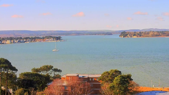 A scenic view of a calm body of water with a sailboat in the foreground. In the distance, there is a shoreline with several colorful buildings and a marina. Lush green trees line the waterfront, and gentle hills are visible on the horizon under a clear blue sky with a few clouds. The overall atmosphere conveys tranquility and natural beauty.