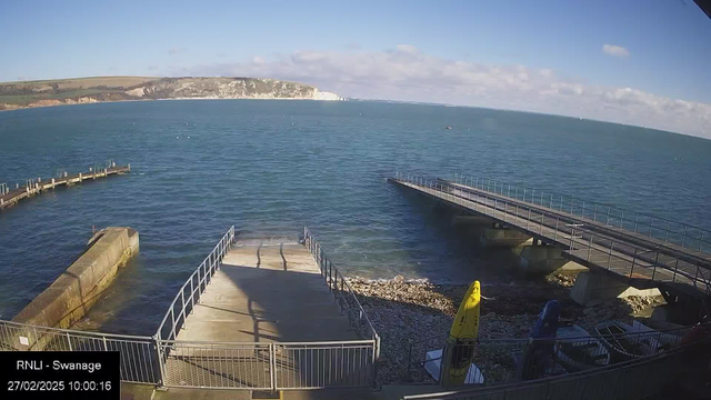 A view of the sea under a clear blue sky. In the foreground, there are two wooden piers extending into the water, one straight and the other curving. To the right, a sloping pathway leads to the water's edge with rocks and a few small boats nearby. A yellow kayak is resting against a railing, and the distant coastline features white cliffs. The time displayed is 10:00:16 on February 27, 2025.