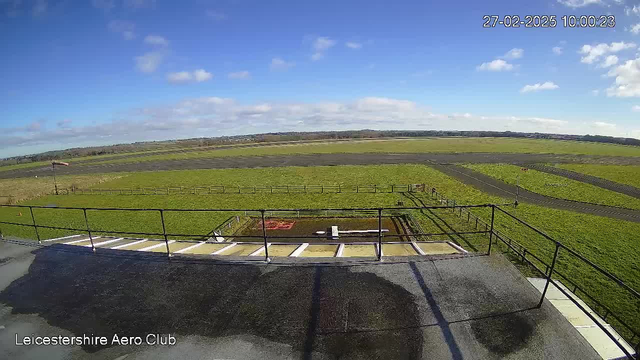 A wide view of a grassy airfield taken from a high vantage point. The sky is mostly clear with a few clouds, and the ground is a mix of green grass and dark asphalt. In the foreground, there is a railing and a concrete platform. The airfield features a runway lined with a fence, and a small area with equipment or structures near the center. In the distance, there are rolling hills and more fields, providing a sense of open space. The image is taken during daylight.