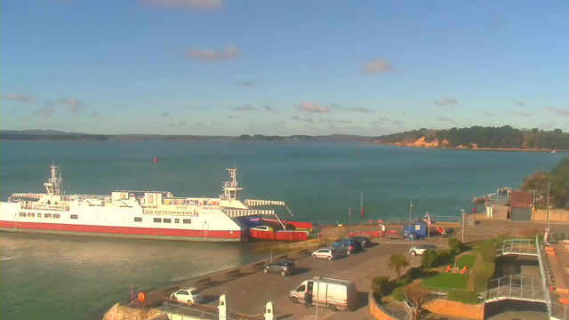 A view of a harbor with a white and red ferry docked at the pier. The water is calm and blue, with a few boats visible in the distance. In the foreground, there is a parking area with several cars and a white van. To the right, a small building and green landscaping can be seen, and in the background, there are green hills and cliffs under a clear blue sky with a few clouds.