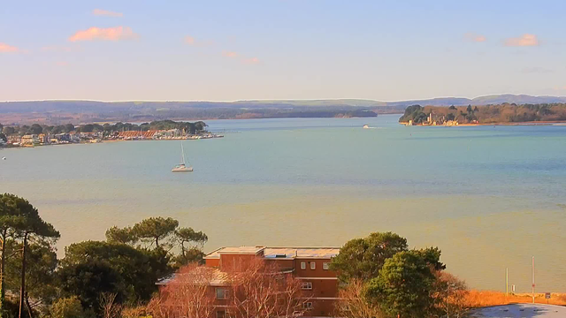 A panoramic view of a calm body of water, with a light blue sky and scattered clouds. In the foreground, there are trees and a low-rise building with reddish walls. Towards the middle of the image, a small sailboat is visible on the water, alongside other boats moored along the shoreline. In the background, there is a marina with several boats, and distant hills or land features can be seen. The scene conveys a peaceful coastal atmosphere.