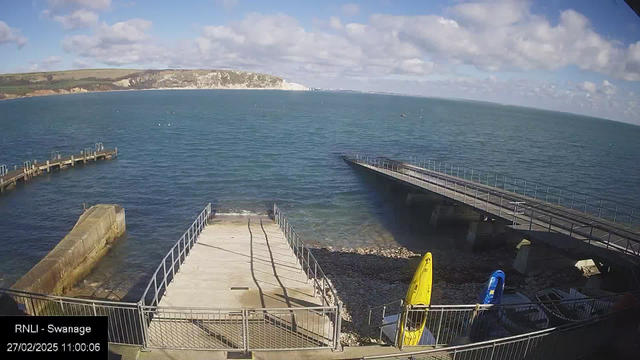 A coastal scene featuring a calm sea under a blue sky with scattered clouds. In the foreground, there is a concrete landing area with a wooden jetty to the left extending into the water. Two colorful kayaks, one yellow and one blue, are resting on the shore beside the landing area. The water is a deep blue, with a rocky beach visible in the lower right corner. In the background, cliffs are visible on the horizon.