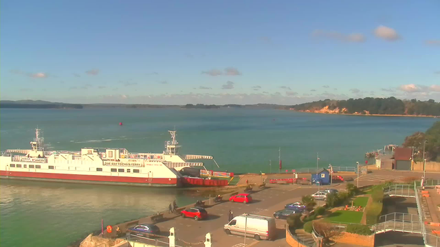 A ferry is docked at a pier, with vehicles parked nearby. The background features a calm blue sea under a sunny sky with scattered clouds. A coastal landscape with green trees rises on the right, while the left side shows a red buoy in the water. The area is bustling with people walking along the dock.